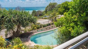 A curved pool with a clear view of the ocean, surrounded by lush greenery and a fenced area.
