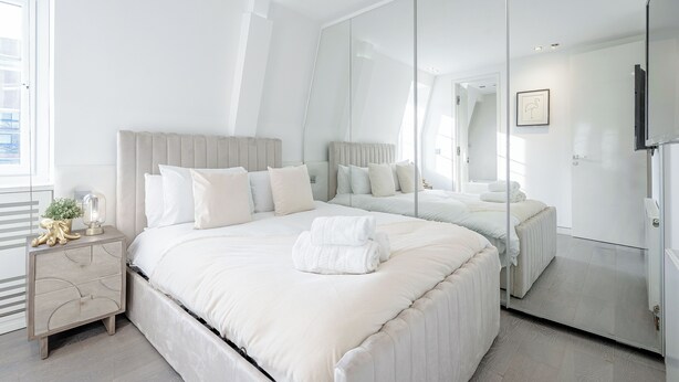 A modern bedroom with a large bed, a bedside table, a mirror, and a view of the city.