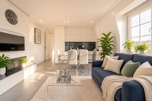 Un salon moderne avec un canapé, une table basse et un téléviseur. On y trouve également un coin repas avec des chaises et une table, ainsi qu’une cuisine à l’arrière-plan.