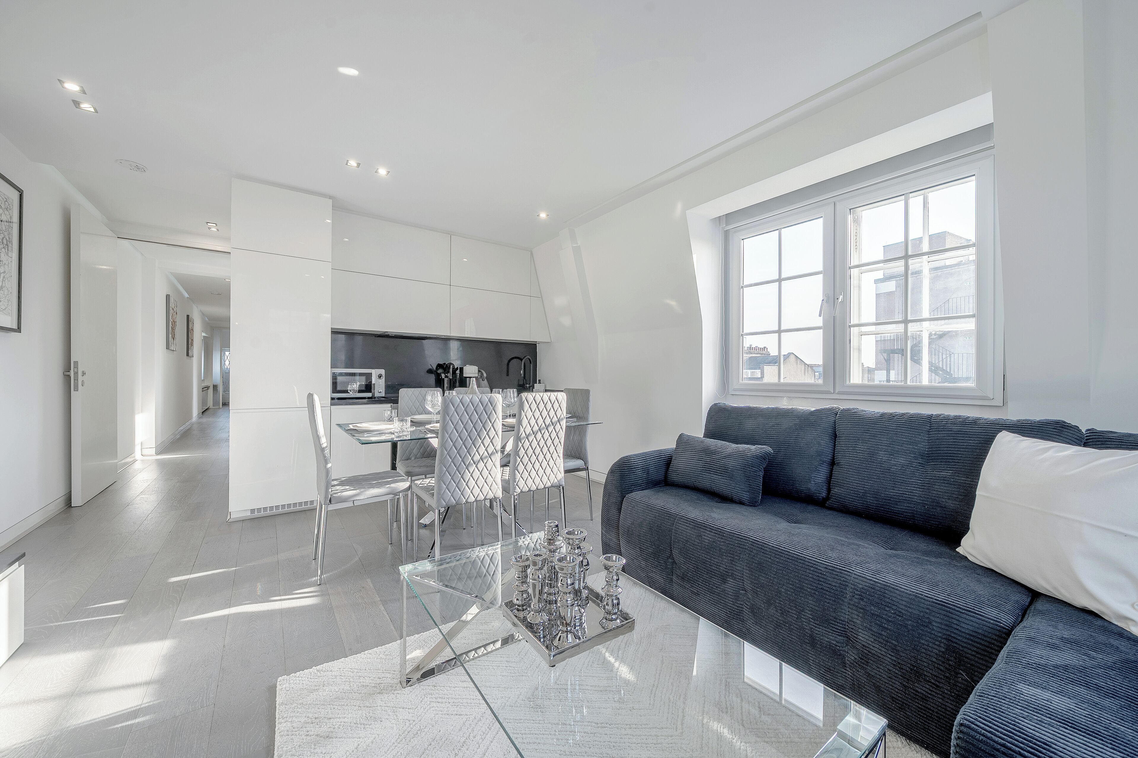Un salon moderne avec un canapé gris foncé, une table basse en verre et un coin repas comprenant une table et des chaises blanches.