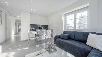 Un salon moderne avec un canapé gris foncé, une table basse en verre et un coin repas comprenant une table et des chaises blanches.