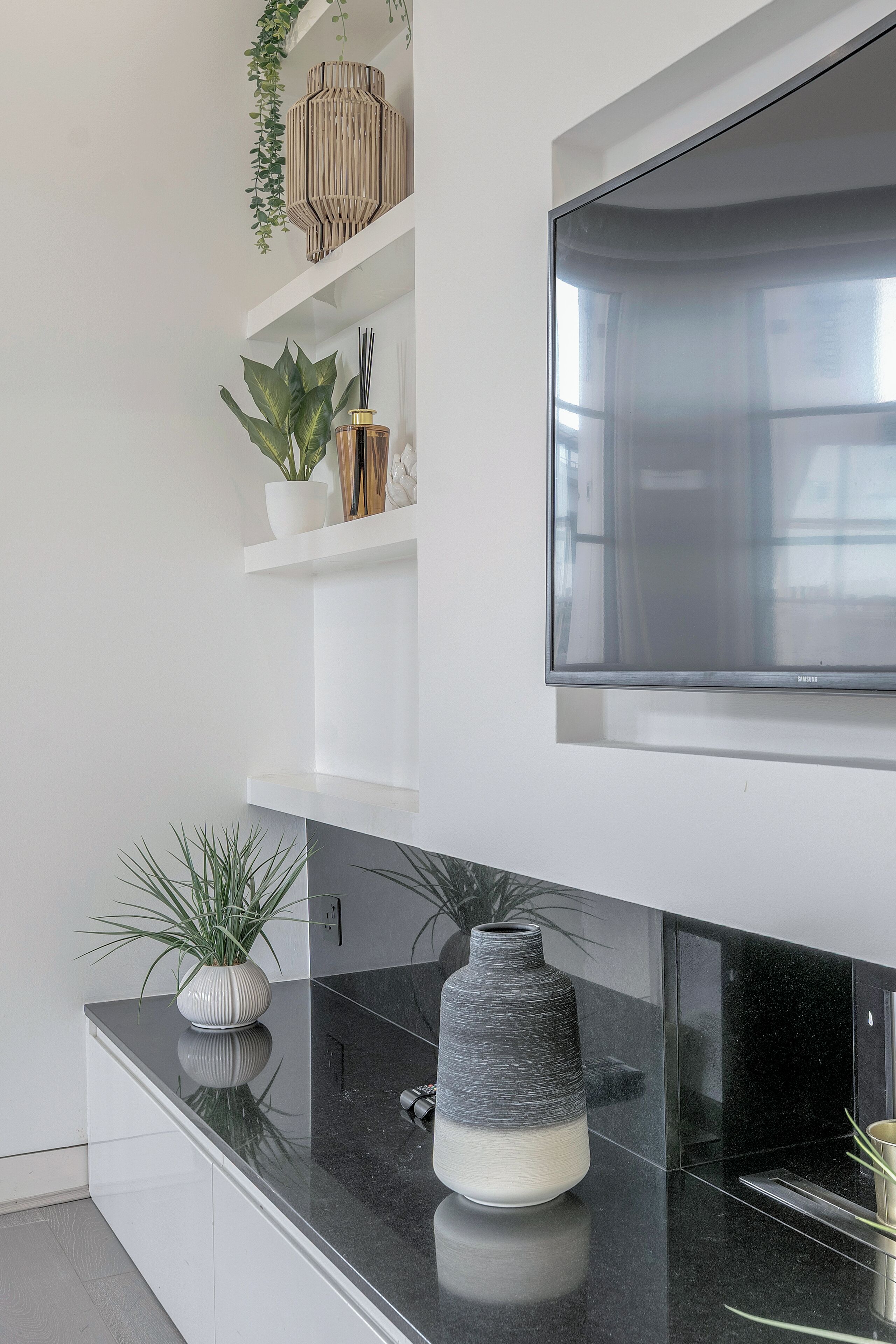 Une cuisine moderne avec un plan de travail noir, des meubles blancs, un grand téléviseur à écran plat fixé au mur et des plantes décoratives disposées sur des étagères.