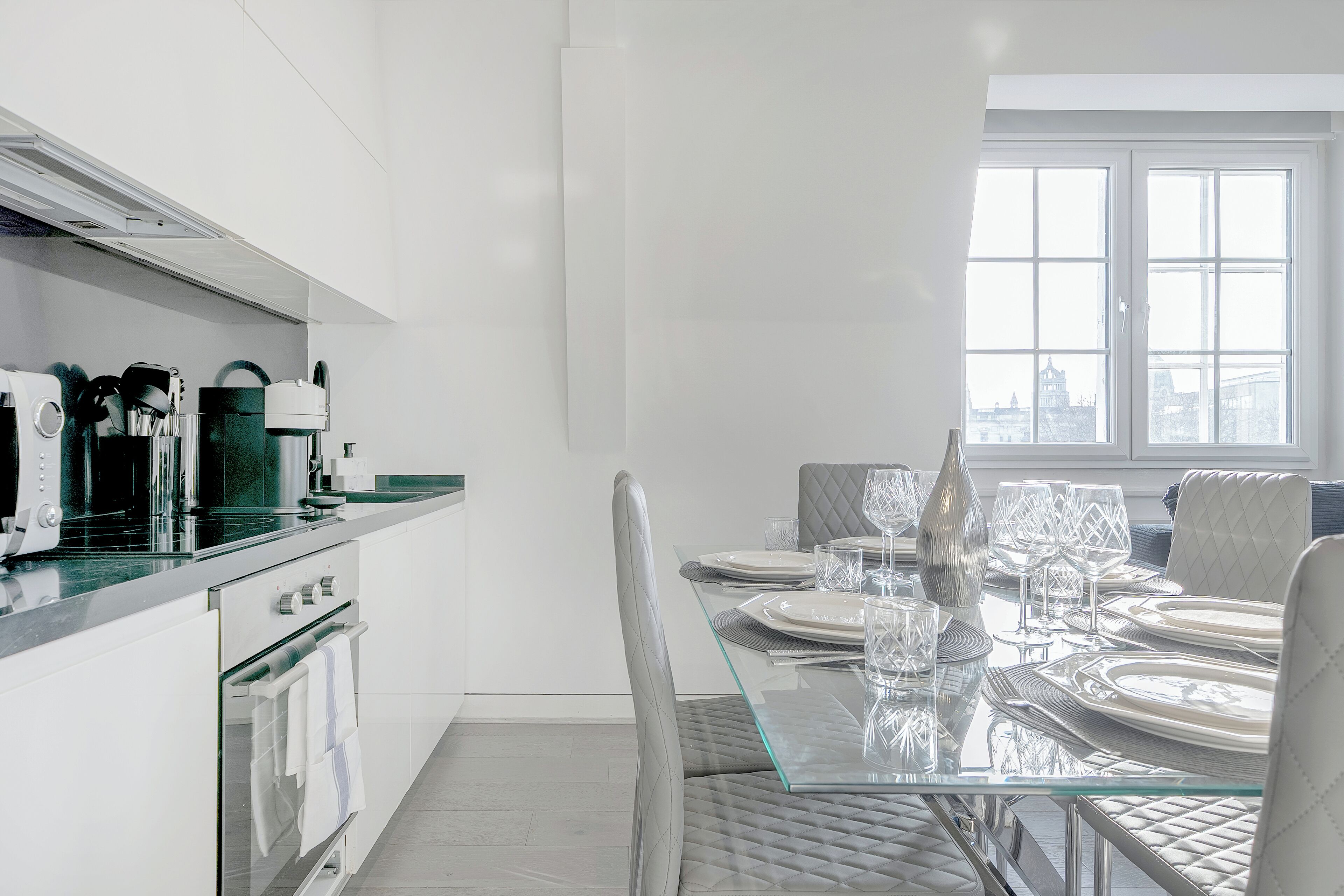 A modern kitchen with a glass dining table set for six, stainless steel appliances, and a city view through large windows.