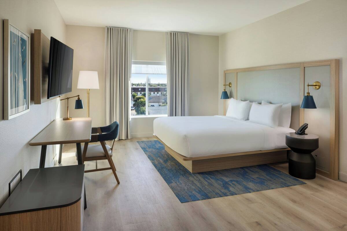 Habitación de hotel con cama, escritorio, silla, televisor, lámparas y vistas al exterior.