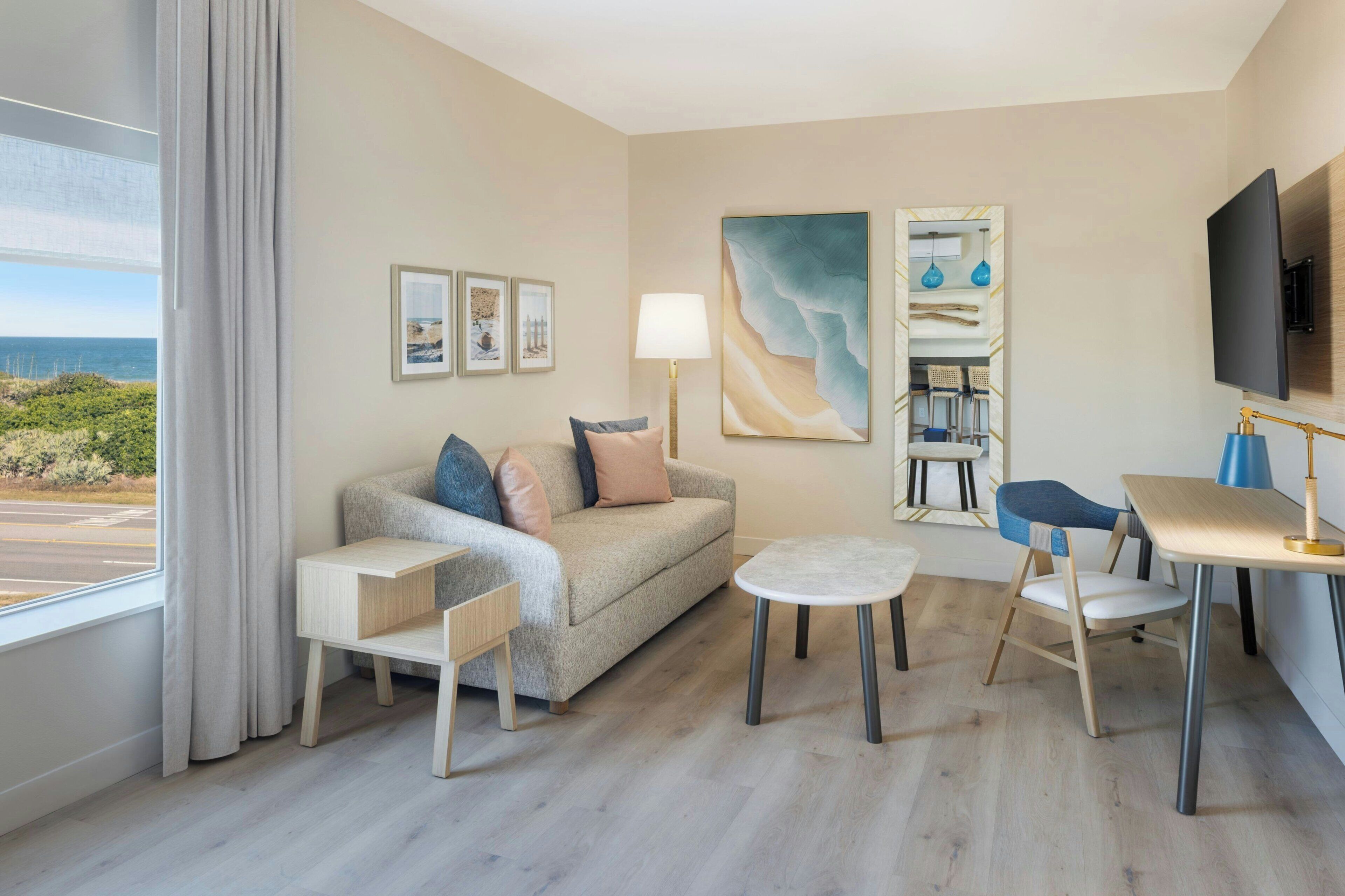 A modern hotel room with a sofa, a small table, a chair, a TV, and a painting.