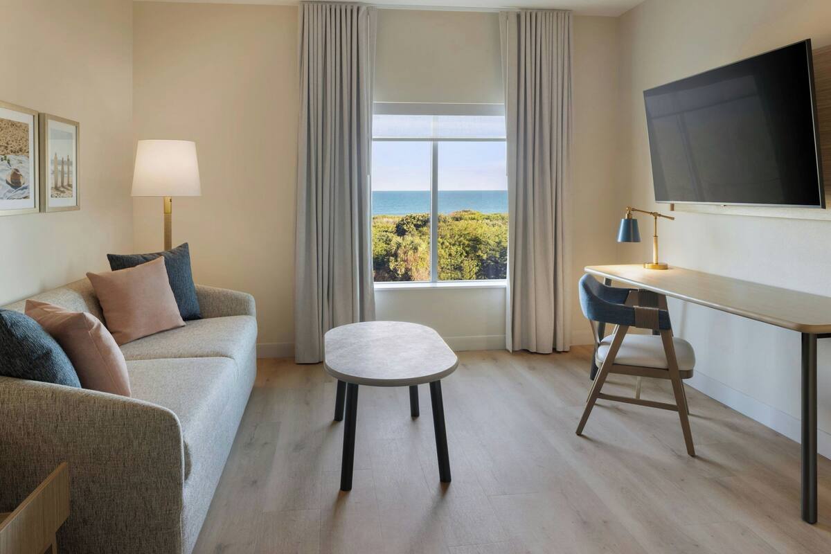 Habitación de hotel con sofá, una mesita, escritorio, silla, televisor y ventana con vistas al mar.
