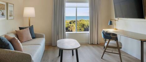 A hotel room with a sofa, a small table, a desk, a chair, a TV, and a window with a view of the sea.