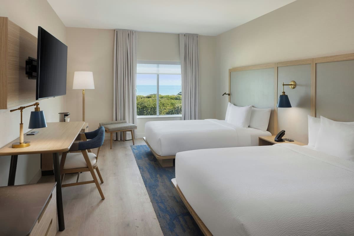 Habitación de hotel con dos camas, un escritorio, una silla, un televisor, una lámpara y vista al mar.