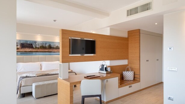 A modern hotel room with a wooden desk, a bed, a TV, and a painting of a landscape.