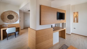 Ein modernes Hotelzimmer mit Schreibtisch aus Holz, einem Sessel, einem an der Wand montierten Fernseher und einem großen Schminkspiegel.