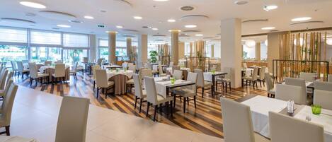 A spacious dining area with multiple tables and chairs, large windows, and a modern ceiling design.