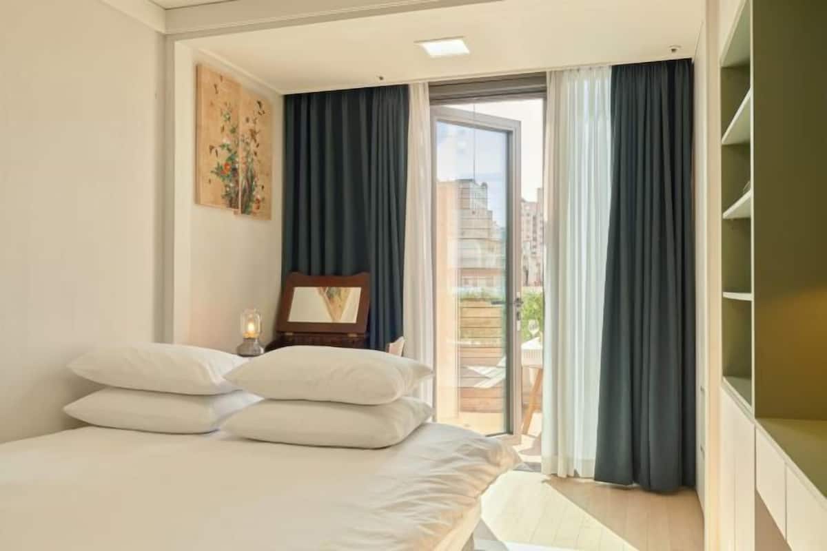 A bedroom with a bed, a nightstand, a painting, and a view of the outdoors.