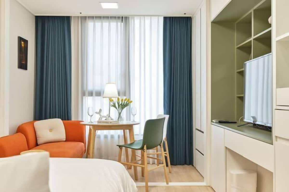 A hotel room with a bed, a desk with a computer, a chair, a small table with a lamp, and a window with curtains.