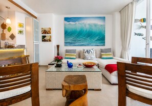 Uma sala de estar com uma grande pintura de ondas, um sofá branco, uma mesa de centro de vidro e cadeiras de madeira.