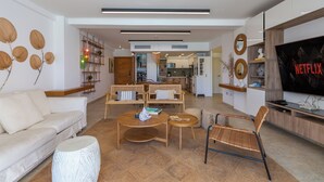 A modern living room with a sofa, coffee table, and chairs, a kitchen in the background, and a TV on the right.