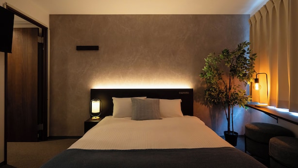 Een hotelkamer met een groot bed, een nachtkastje met lamp, een potplant en uitzicht op het buitenleven.