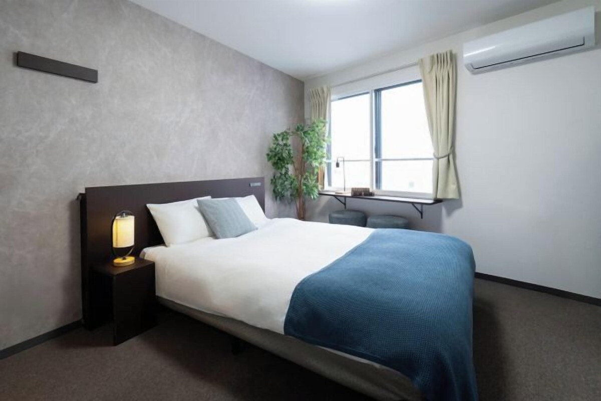 A modern hotel room with a large bed, a bedside table with a lamp, a potted plant, and a window with curtains.