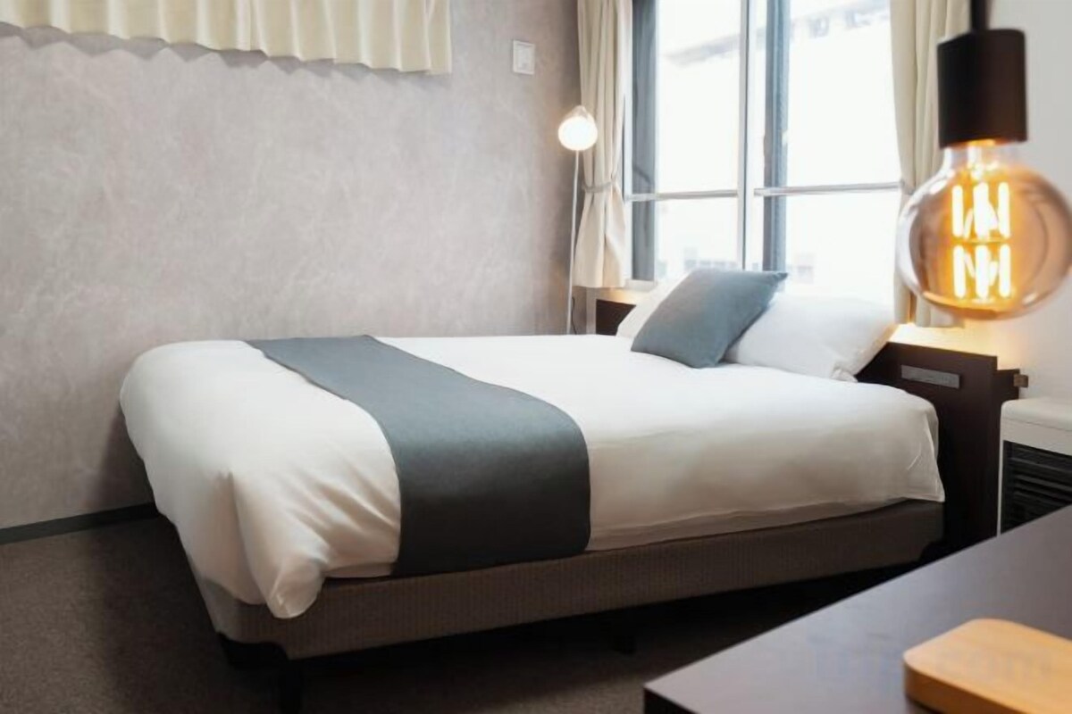 Una cama bien tendida con una colcha blanca y gris, una lámpara de noche y una ventana con cortinas.