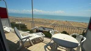A balcony with white plastic chairs and a table, overlooking a beach with rows of umbrellas and people. - Seaside Bliss apartment with front sea view (Lignano Sabbiadoro)