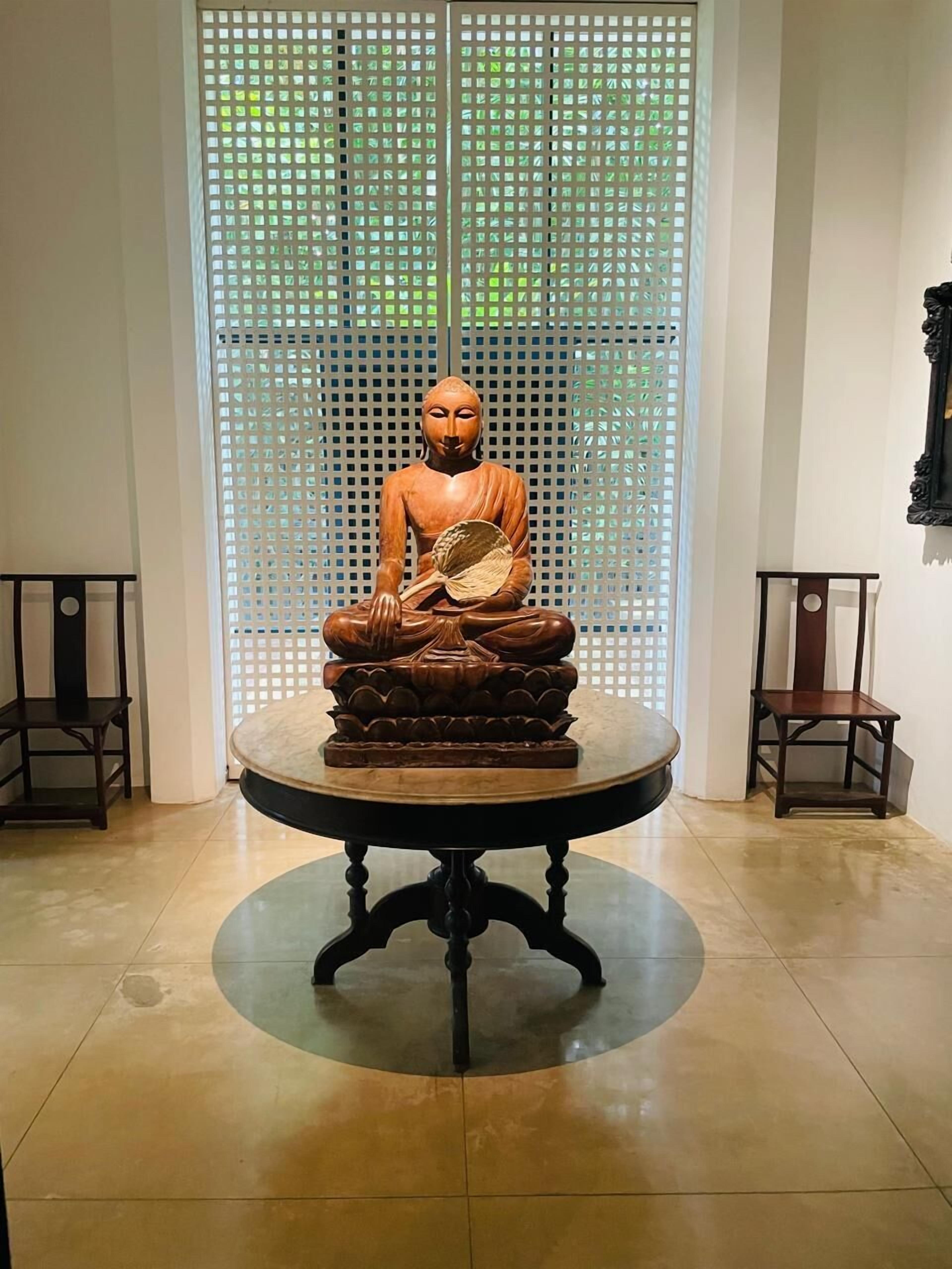 A statue of a seated figure on a pedestal in a room with a tiled floor and a window with a lattice design.