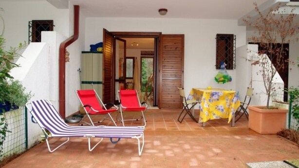 Un espace patio comprenant deux chaises longues à rayures, une chaise rouge, une petite table recouverte d’une nappe à motifs floraux jaunes et un pot de fleurs.