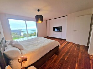 A bedroom with a large bed, a bedside table, a hanging light fixture, a TV, and a sliding door to a balcony.