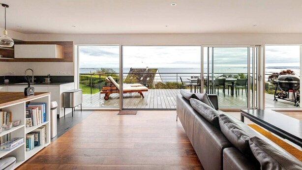 A modern kitchen with a dining area, a sofa, and a balcony with a view of the sea.