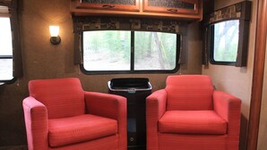 Een compact camperinterieur met twee rode stoelen, een klein zwart tafeltje en een raam met uitzicht op de omgeving.
