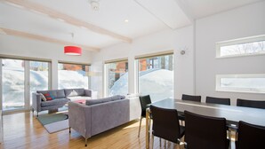 Una sala de estar moderna con un sofá gris, una mesa de comedor con sillas, ventanas amplias con nieve visible, y una lámpara colgante roja.