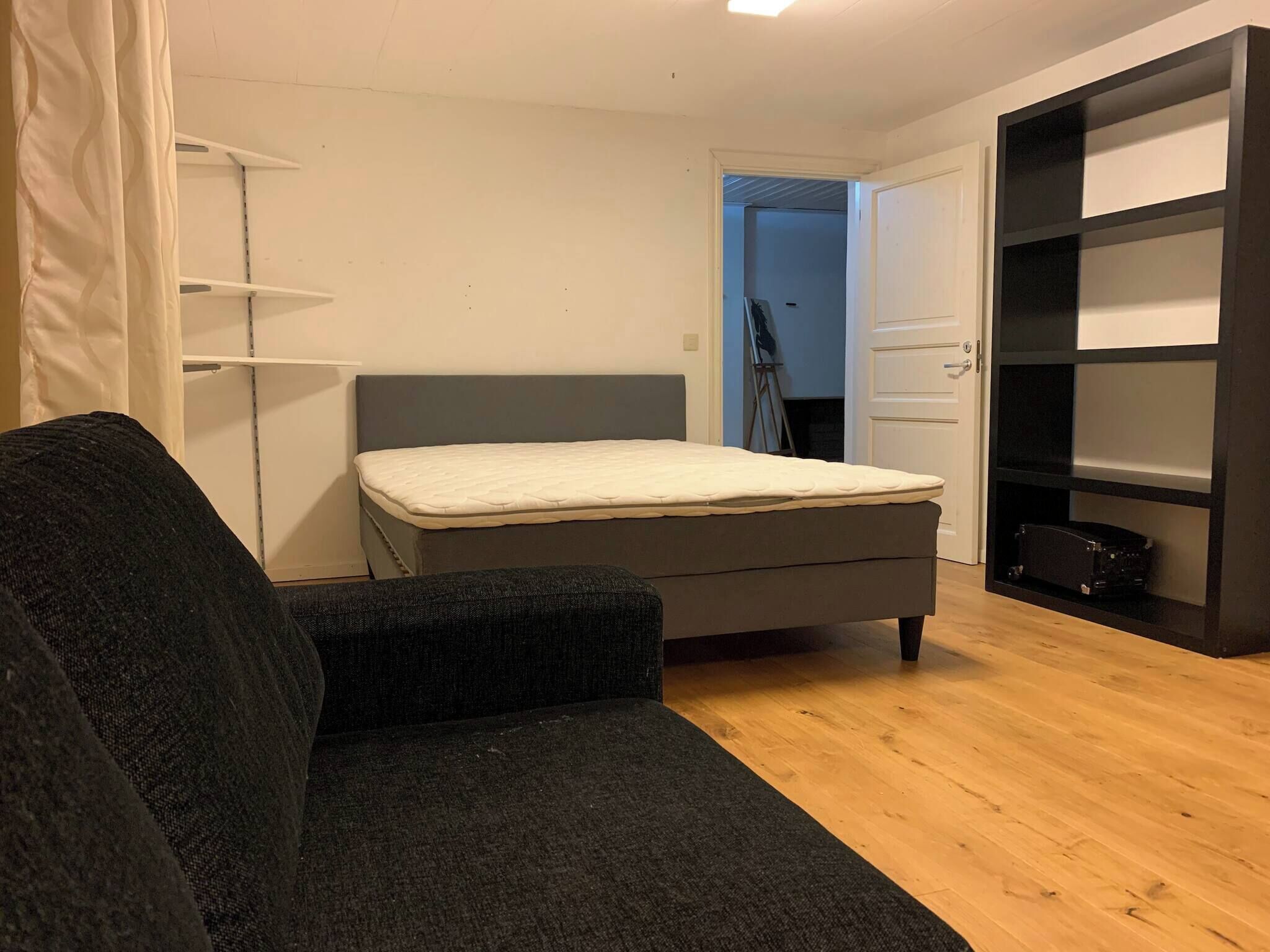 A small, modern bedroom with a grey sofa, a single bed, a wardrobe, and a door leading to another room.