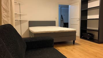 Um quarto pequeno e moderno, com um sofá cinzento, uma cama de solteiro, um guarda-roupa e uma porta que dá para outro espaço.