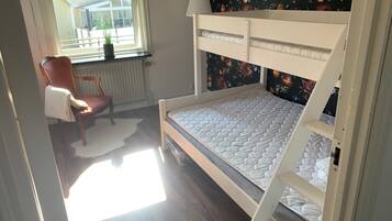 Uma cama beliche com colchão, uma cadeira, um radiador e uma janela com vista para edifícios.