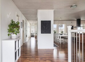 A spacious hallway with wooden flooring, white walls, and a central column. There is a dining area with white chairs and a table, and a potted plant on the left. - Ronneby Torneryd Retreat (Ronneby)