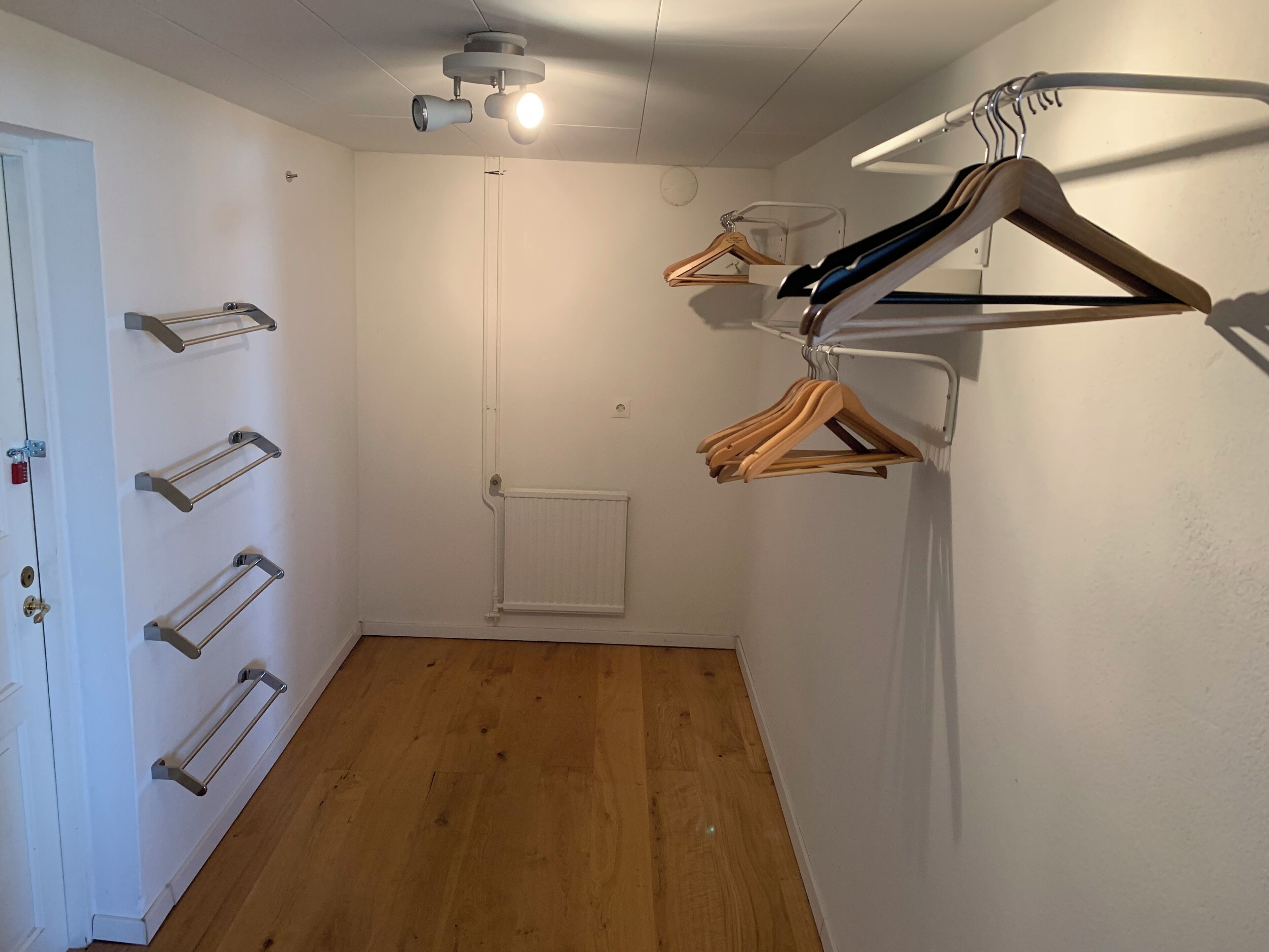 Una habitación con suelo de madera, un radiador blanco, una puerta con cerradura y un proyector instalado en el techo.