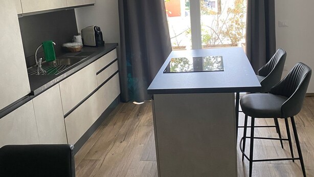 Moderni keittiö, jossa on keskeinen saareke, ruokailutila kahdella tuolilla sekä suuret ikkunat, joista on näkymä ulos.