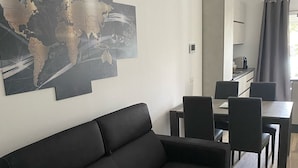 Moderni olohuone, jossa on musta sohva, ruokailutila pöytineen ja tuoleineen sekä taustalla keittiö.