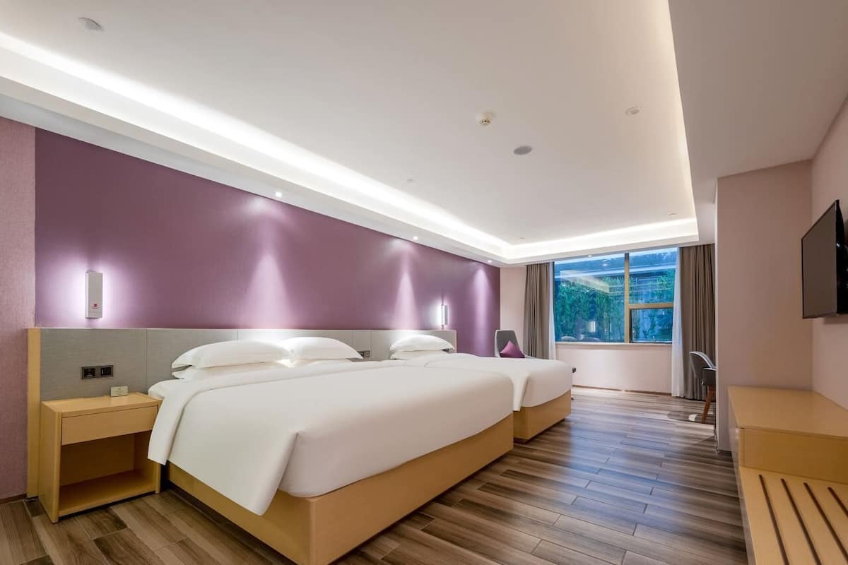 A modern hotel room with a large bed, a bedside table, a TV, and a window with a view of greenery.