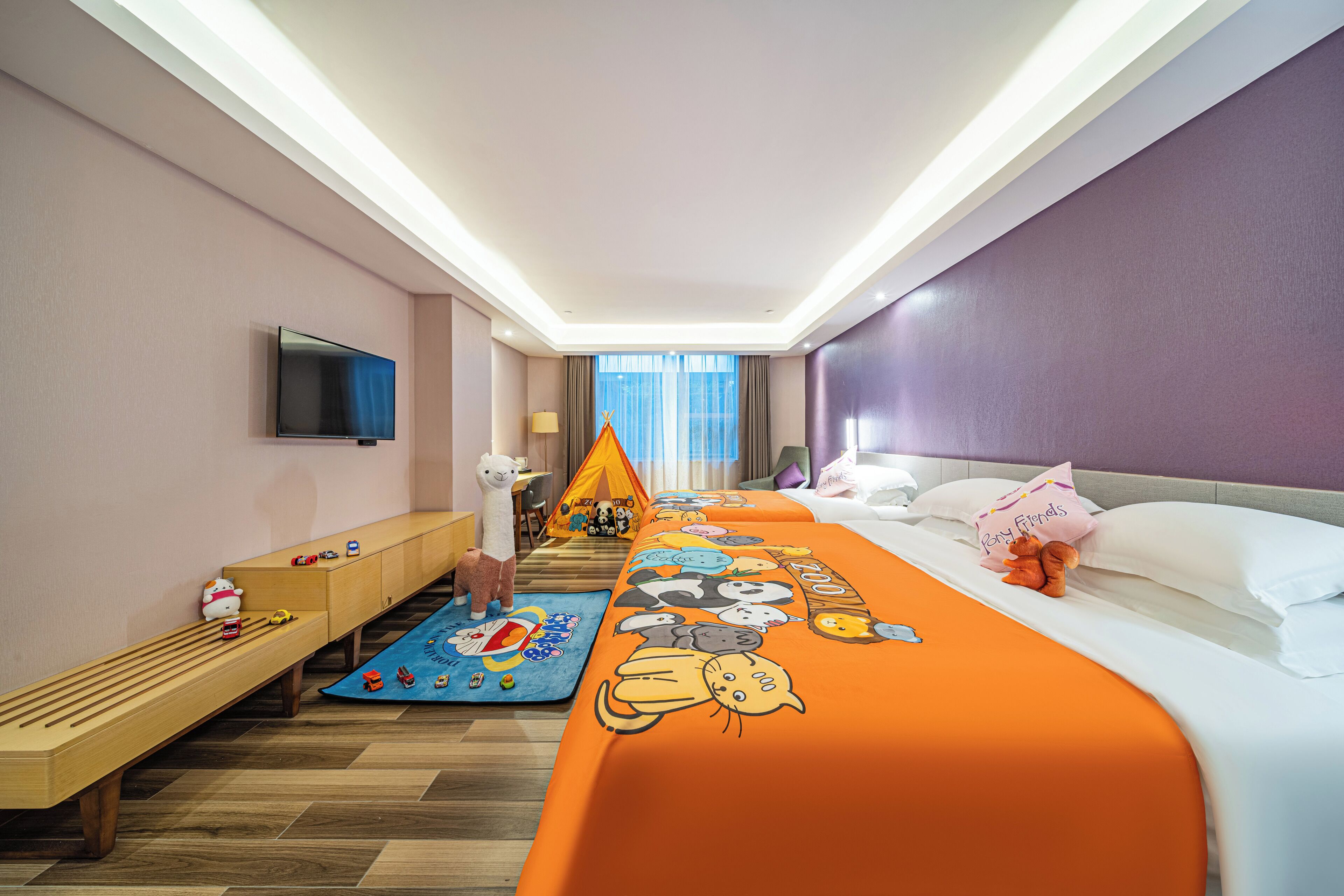 A hotel room with a bed, a TV, a bench, a play mat, and a window with curtains.