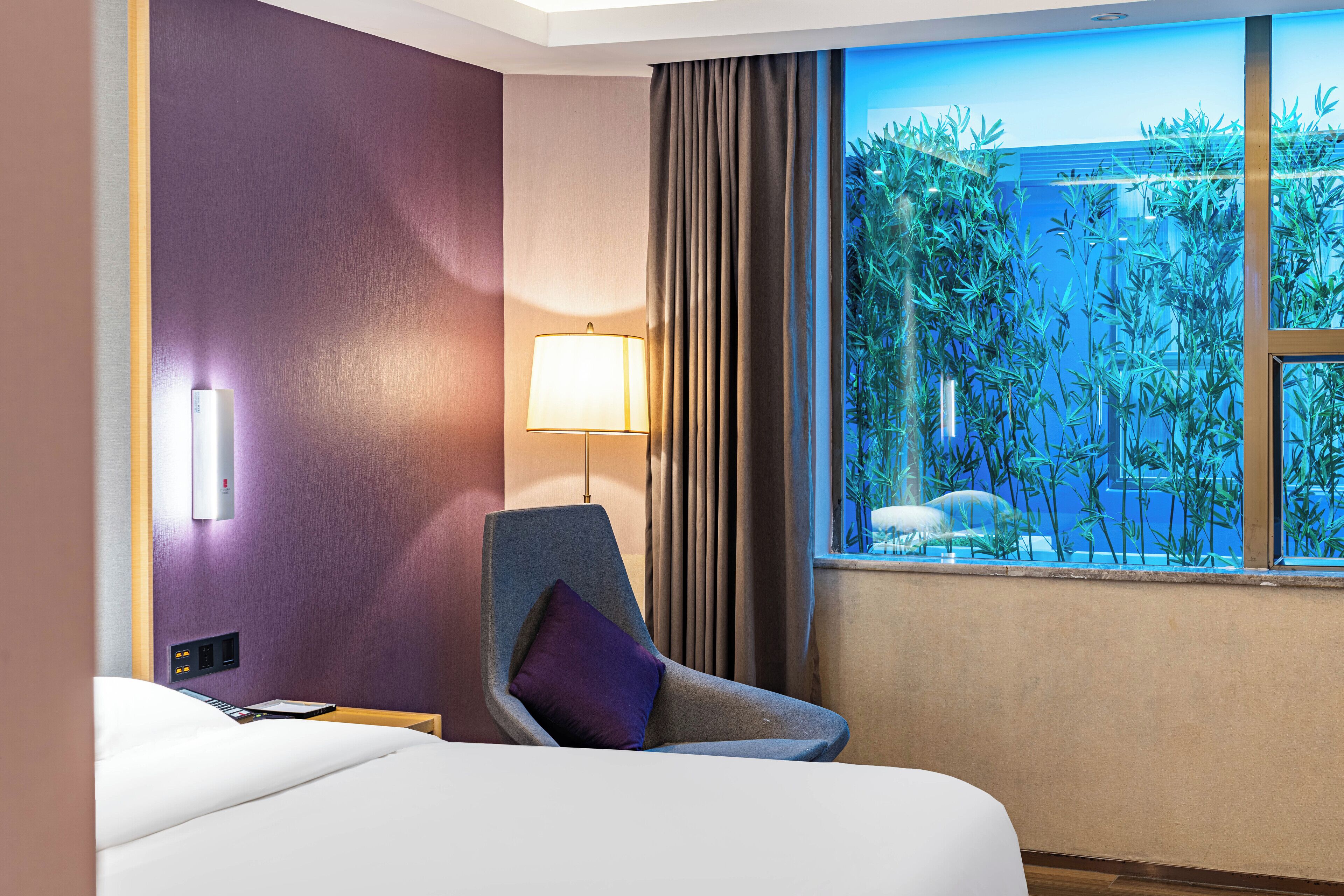 A hotel room with a bed, a chair, a lamp, and a window with a view of greenery.