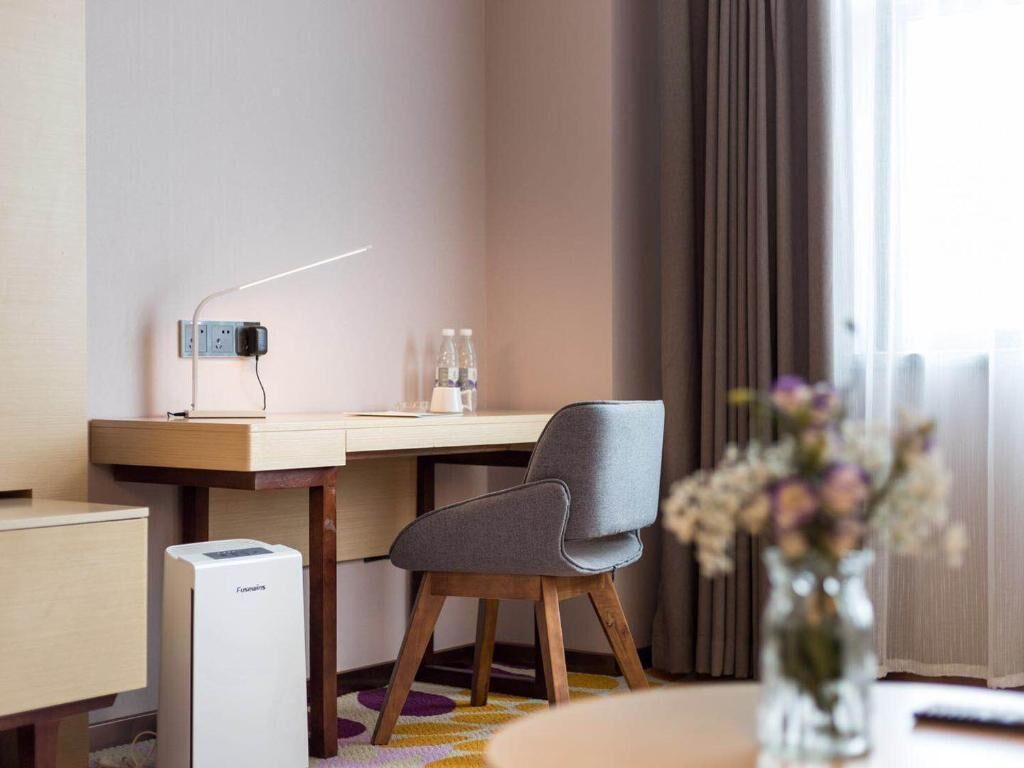 Una habitación de hotel moderna con un escritorio de madera, una silla gris, un purificador de aire compacto y un jarrón con flores.