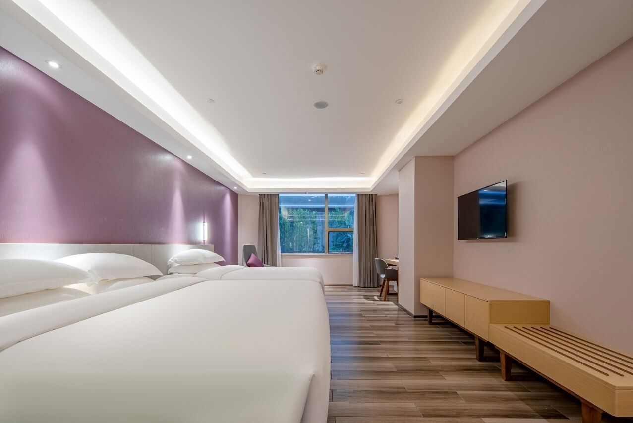 A modern hotel room with a large bed, a desk, a bench, and a TV on the wall.