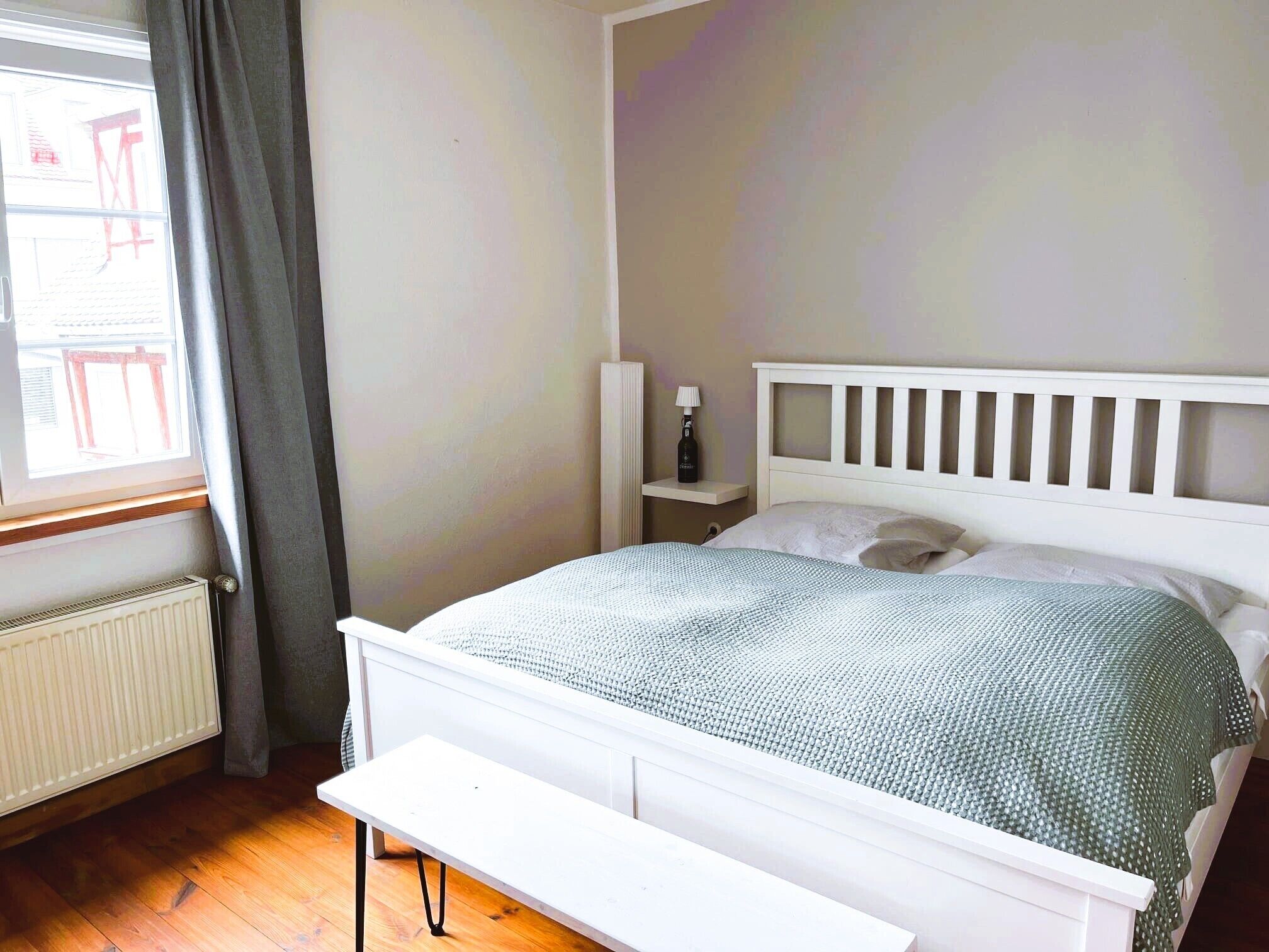Une chambre à coucher avec un lit blanc, une table de chevet, une lampe et une fenêtre avec des rideaux.