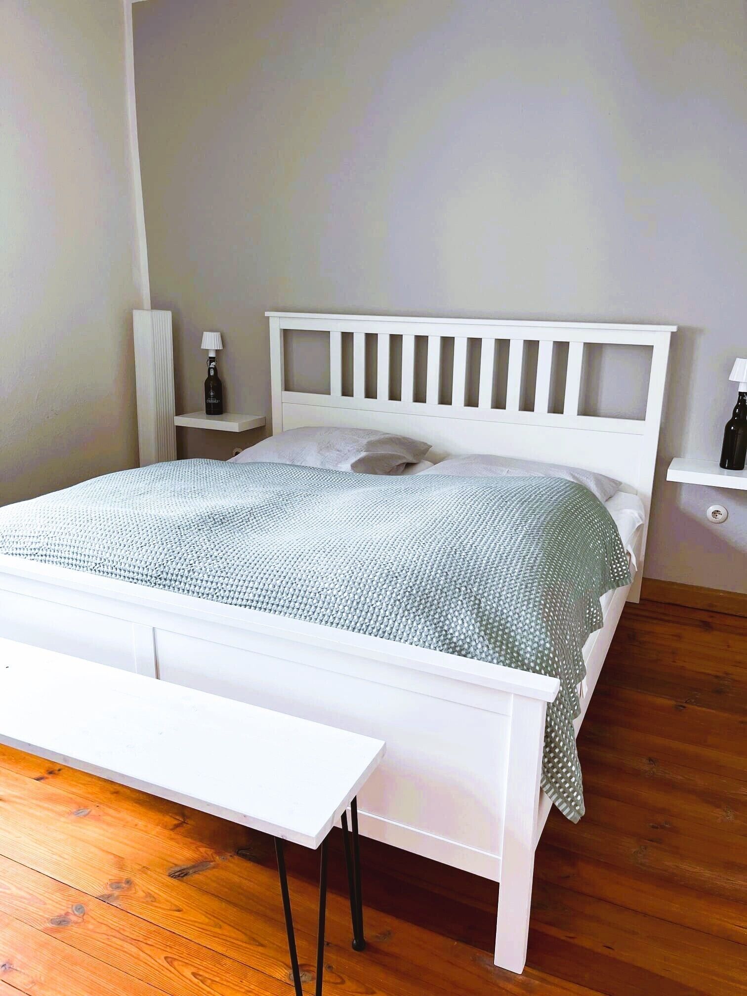 Ein ordentlich eingerichtetes Schlafzimmer mit einem weißen Bett, einem Nachttisch, einer Lampe und einem kleinen Regal.