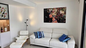 A modern living room with a sofa, a white chair, and a coffee table. There is a painting on the wall and a shelf with bottles.