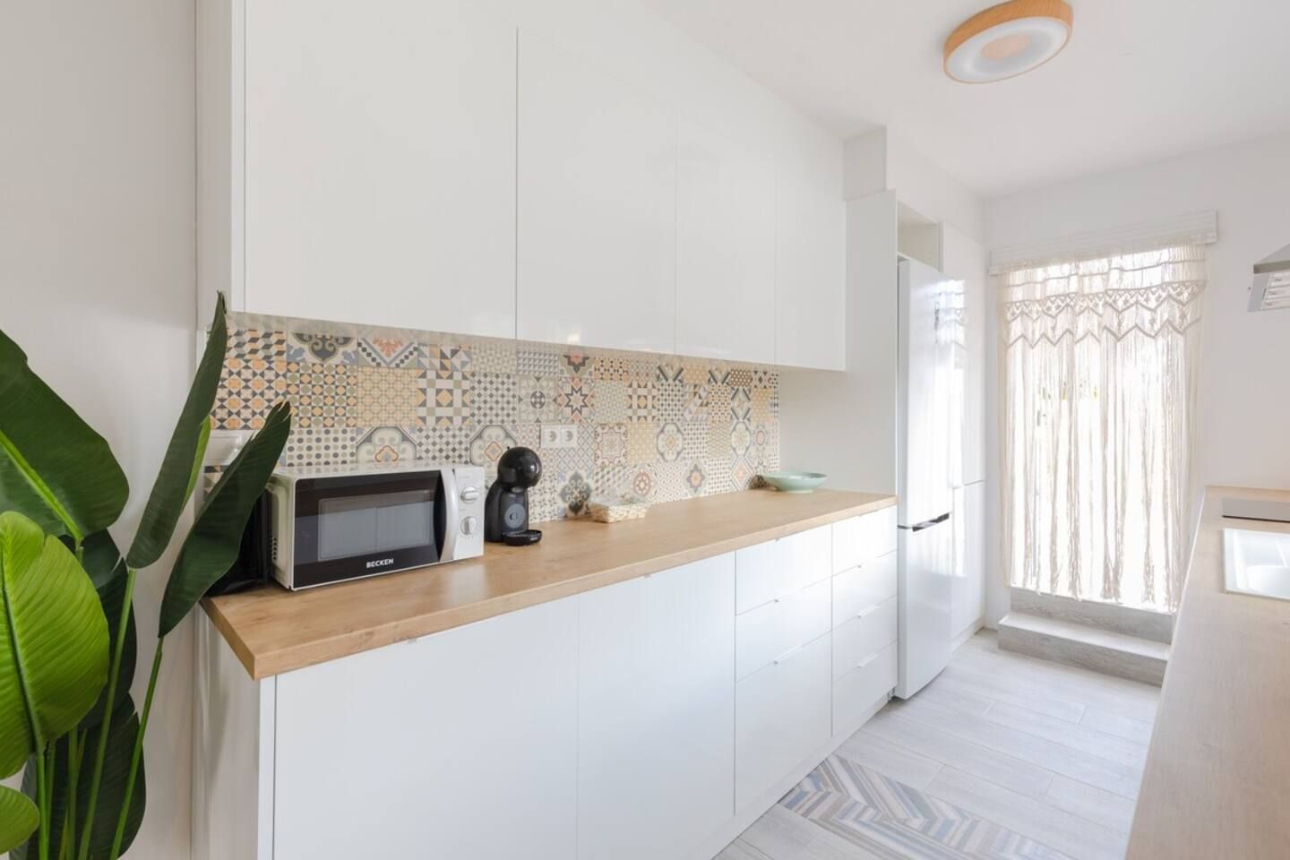 Une cuisine moderne avec des meubles blancs, un pare-feu décoratif, un four à micro-ondes, une machine à café et une fenêtre avec des rideaux.