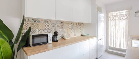Une cuisine moderne avec des meubles blancs, un pare-feu décoratif, un four à micro-ondes, une machine à café et une fenêtre avec des rideaux.