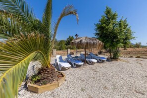 A beachside lounge area with sun loungers and a thatched umbrella, surrounded by gravel and palm trees. - Rustic Escape by Fidalsa (Ibi)