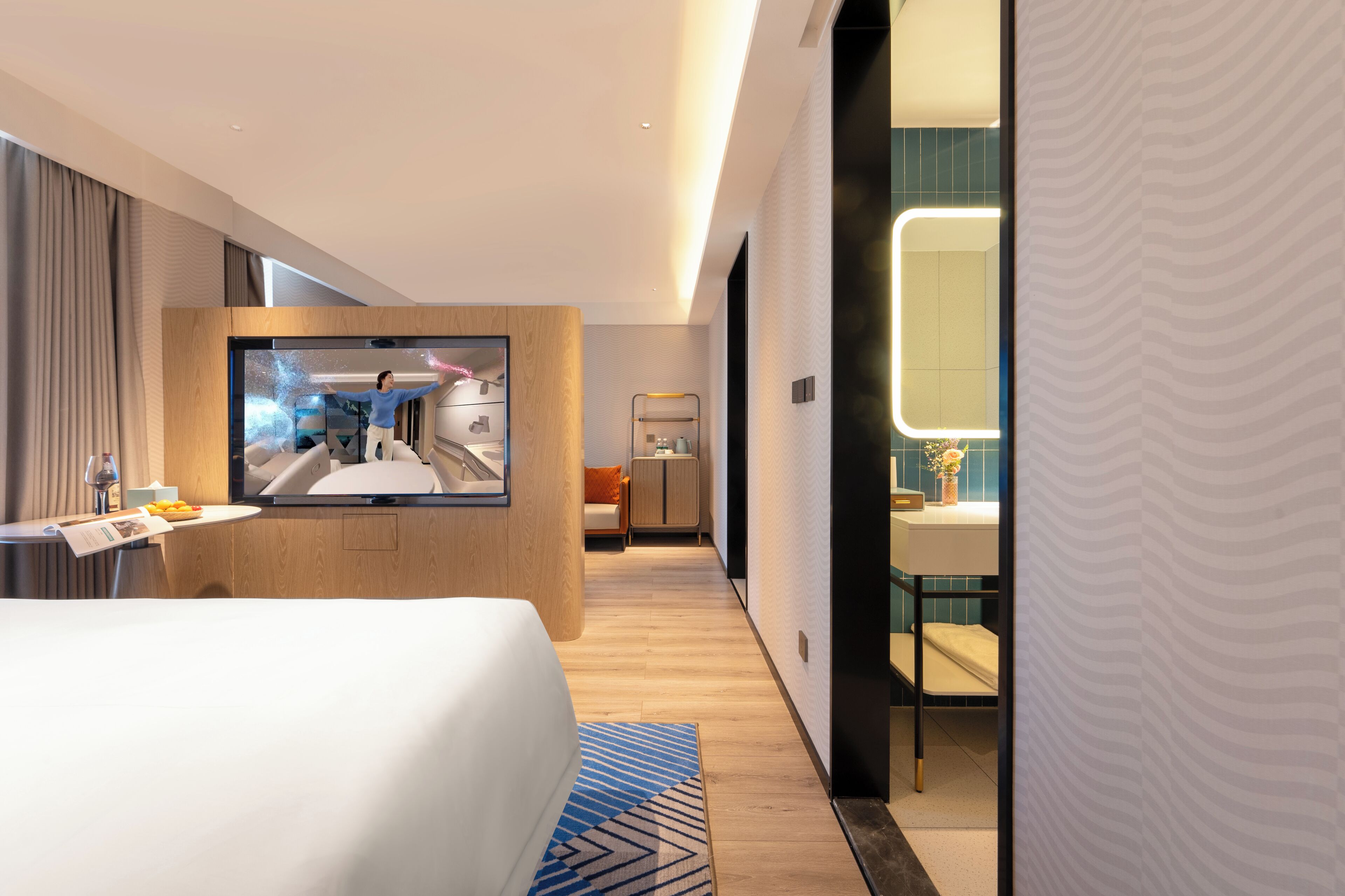 Une chambre d’hôtel moderne dotée d’un grand lit, d’un téléviseur fixé sur un panneau en bois, d’une salle de bain avec un miroir et un lavabo, et d’un couloir menant à une autre chambre.