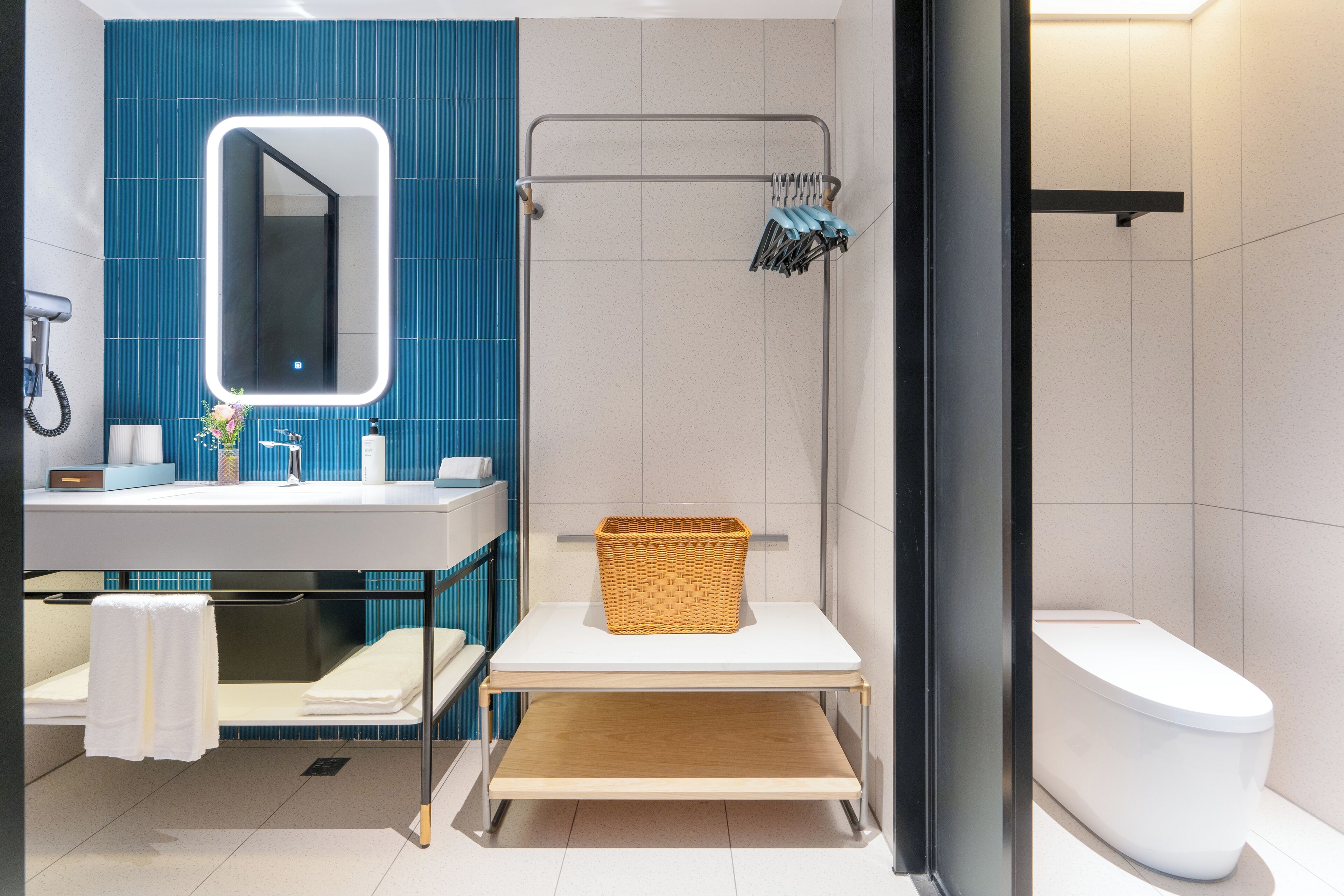 A modern bathroom with a blue tiled wall, a white sink with a mirror, a shower with a towel rack, and a white toilet.