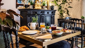 Una mesa de desayuno dispuesta con croissants, fruta y bebidas, rodeada de sillas y un mueble de madera oscura con adornos.